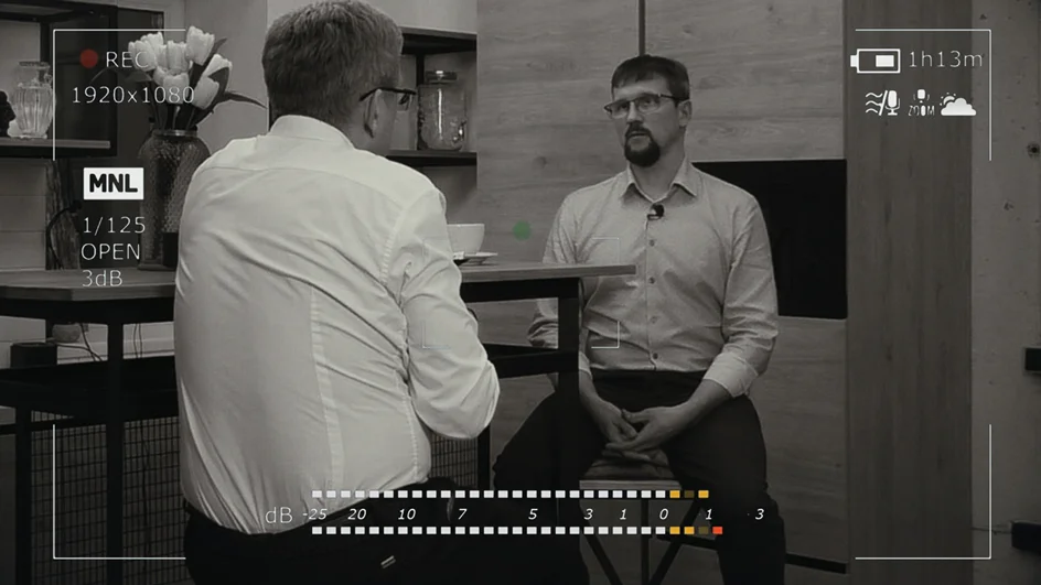 A black and white still from a video showing two men in a discussion.